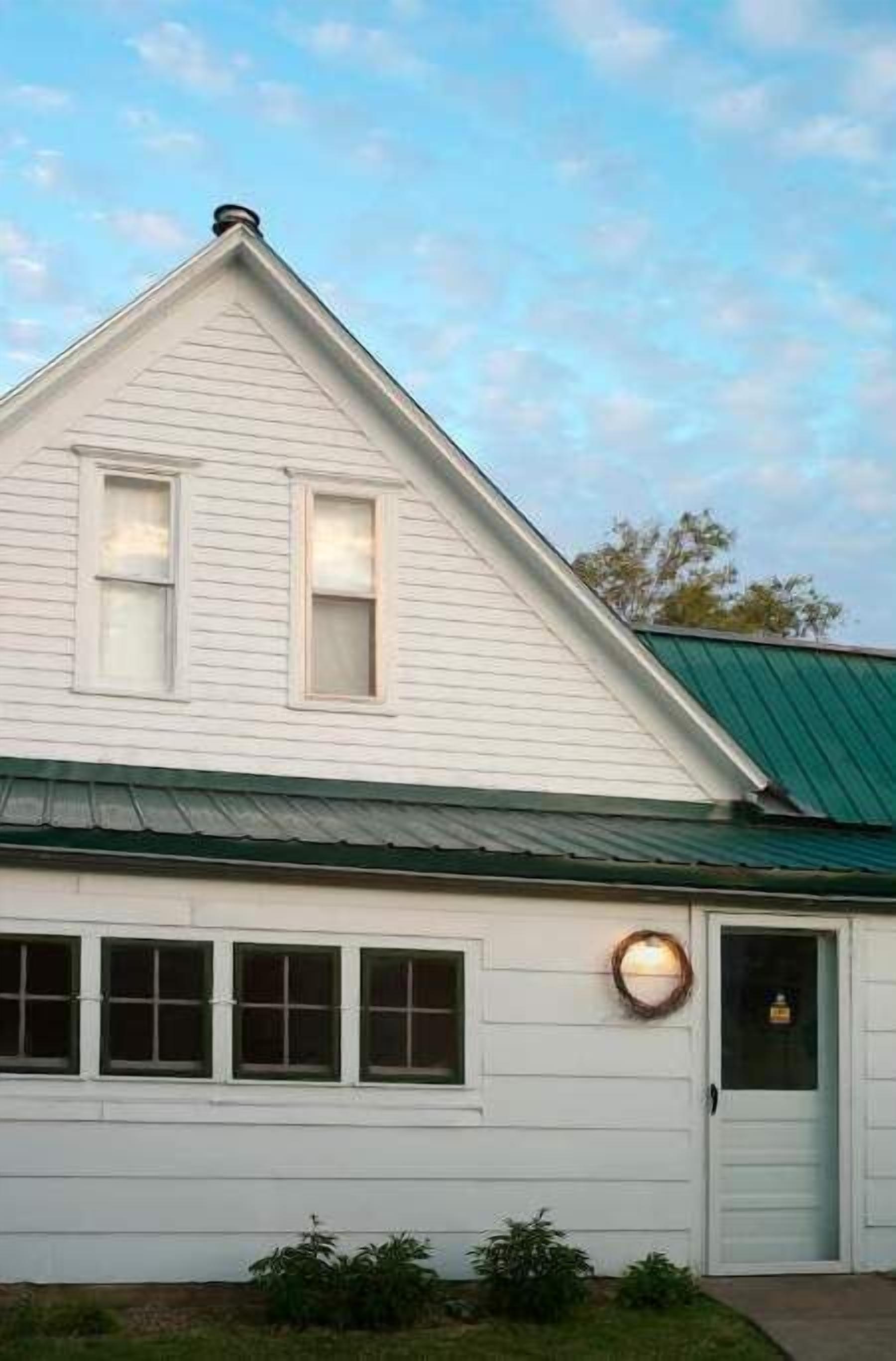 A white building with a green roof, a front door, and a lit wall-mounted light fixture.