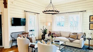 A cozy living room with a wooden ceiling, a large window, a sofa, two armchairs, a coffee table, and a TV.