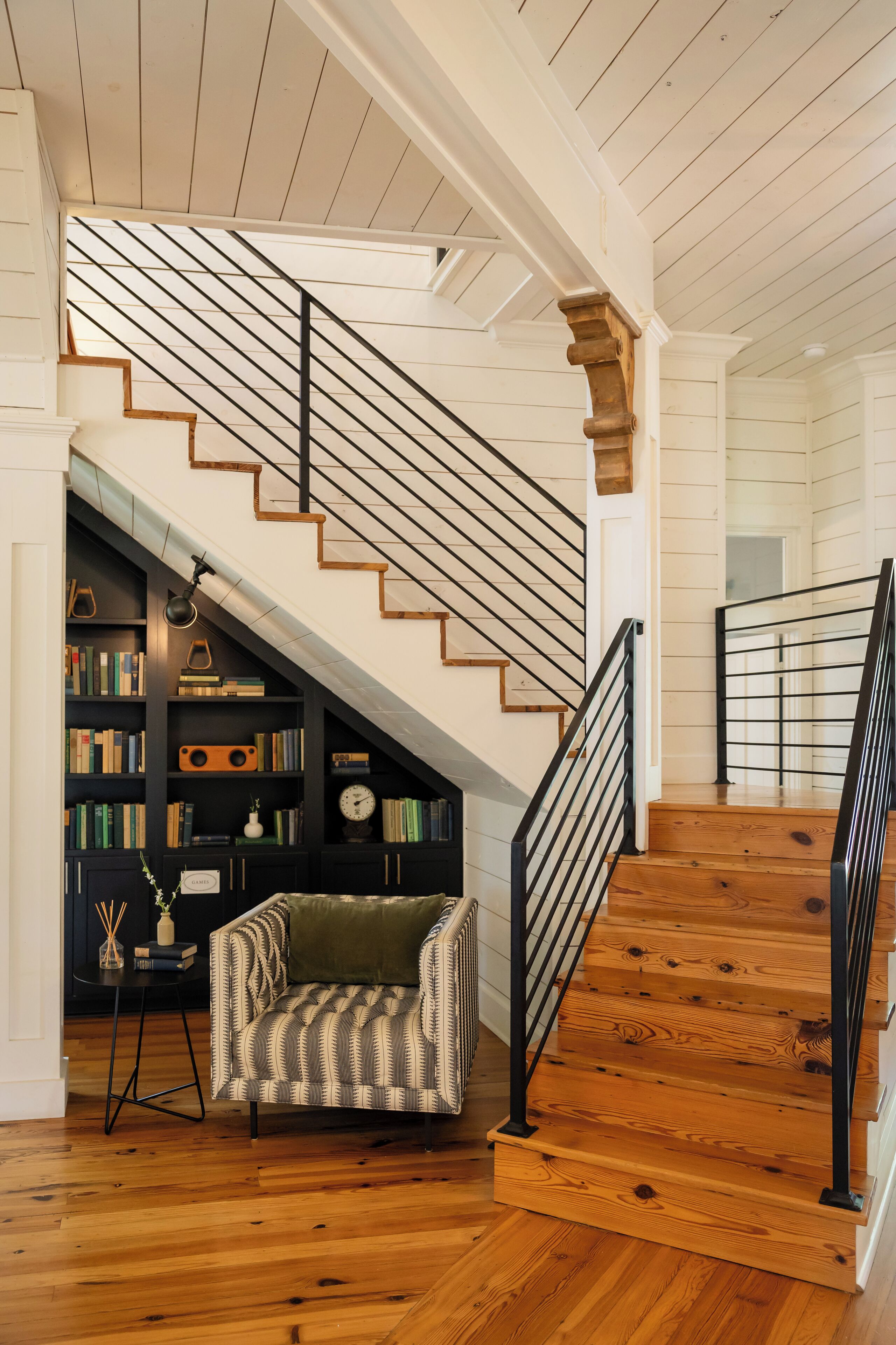 A wooden staircase with black railings, a bookshelf with books and decorative items, and a patterned armchair.