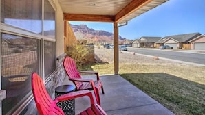 A covered porch with red Adirondack chairs and a small table, overlooking a street with houses and a mountain range. - Moab Oasis among the Redrocks! (Moab)