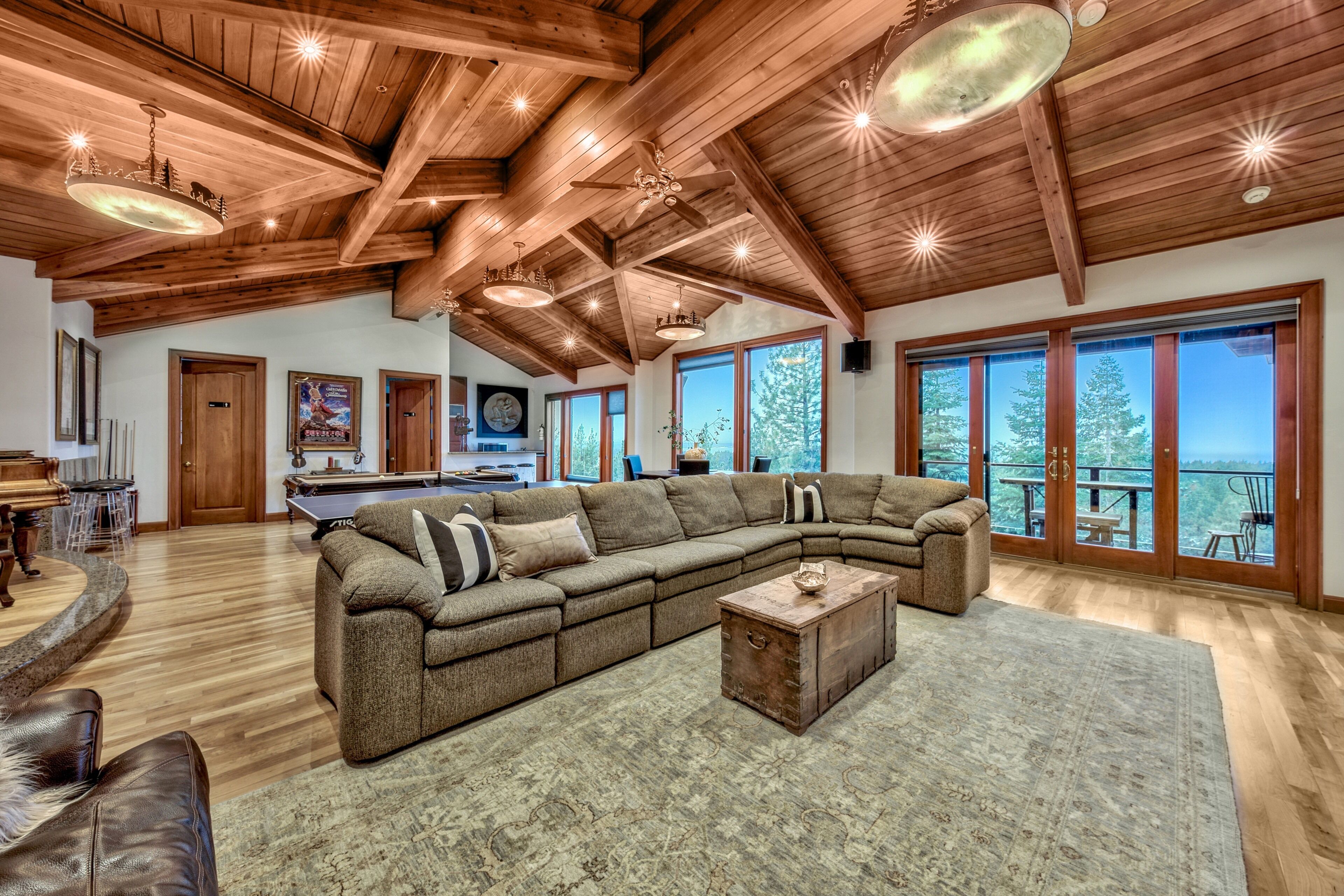 A spacious living room with a large sectional sofa, wooden ceiling beams, and large windows offering a view of the outdoors.