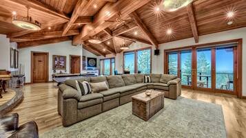 A spacious living room with a large sectional sofa, wooden ceiling beams, and large windows offering a view of the outdoors.