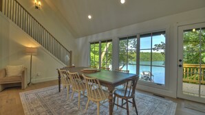 Un coin repas avec une table et des chaises en bois, un escalier et de grandes fenêtres donnant sur un lac.