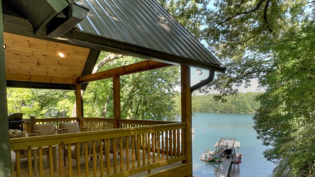Une terrasse en bois avec des sièges extérieurs, donnant sur un lac, un quai et un petit bateau.