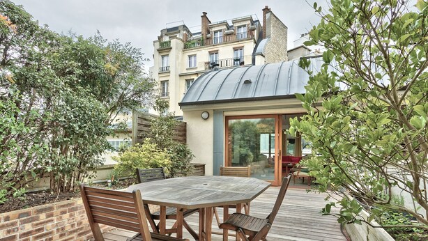 A wooden deck with a table and chairs, surrounded by greenery and a building with a metal roof in the background. - Appartement Dombasle, Tour Eiffel - 3 étoiles - Welkeys (Paris)