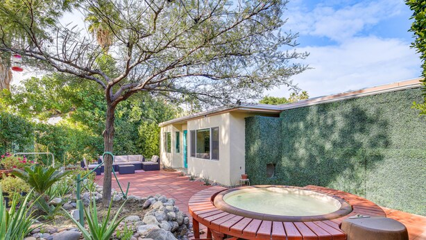 Un jacuzzi con cubierta de madera, rodeado de rocas y plantas, en un patio trasero con una casa y un árbol.