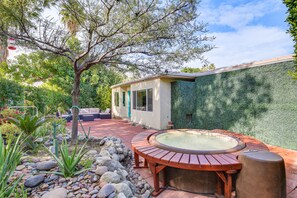 A hot tub with a wooden deck, surrounded by rocks and plants, in a backyard with a house and a tree. - Palm Springs Studio w/ Outdoor Oasis & Hot Tub! (Palm Springs)