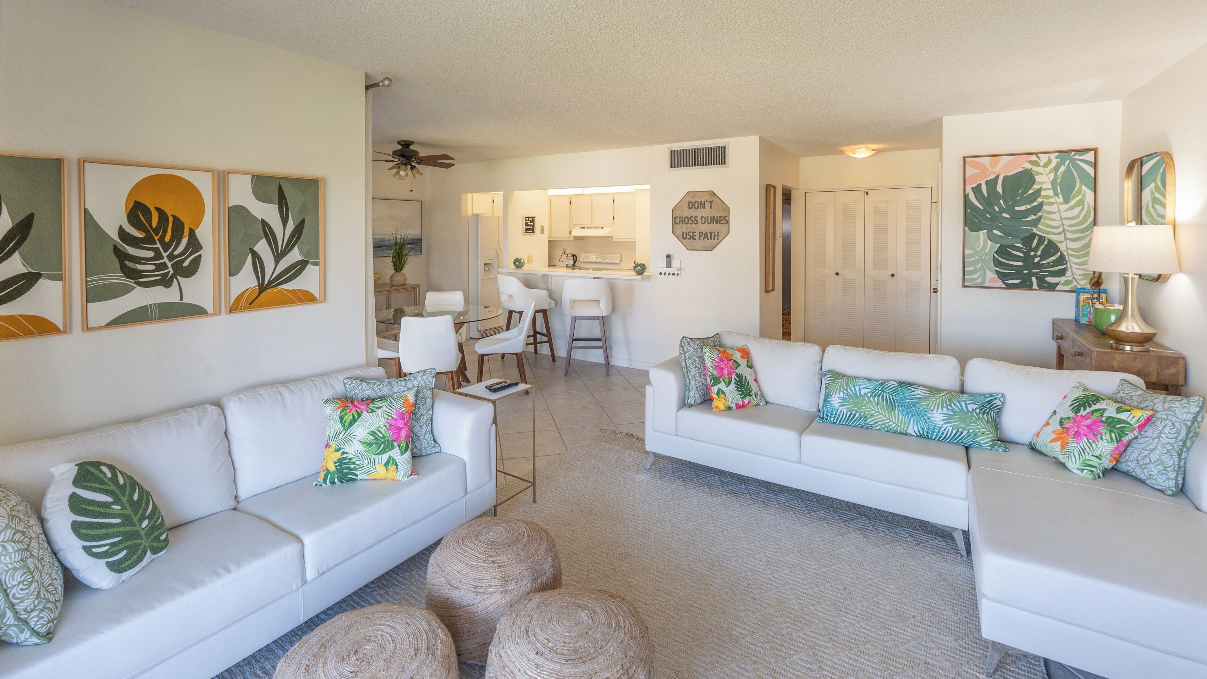 A modern living room with white sofas, tropical-themed cushions, and a kitchen in the background.
