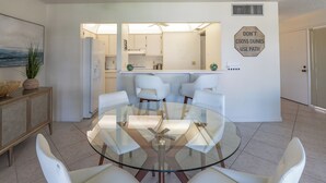 Un comedor moderno con una mesa de vidrio y sillas blancas, una cocina al fondo, y un letrero en la pared que dice 'NO CRUZAR DÍNAS, USE SENDERO'.