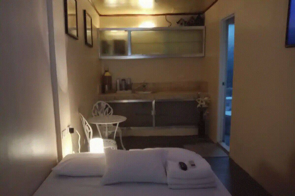 A small, dimly lit room with a bed, a small table, and a chair. There is a window with a view of the outside.