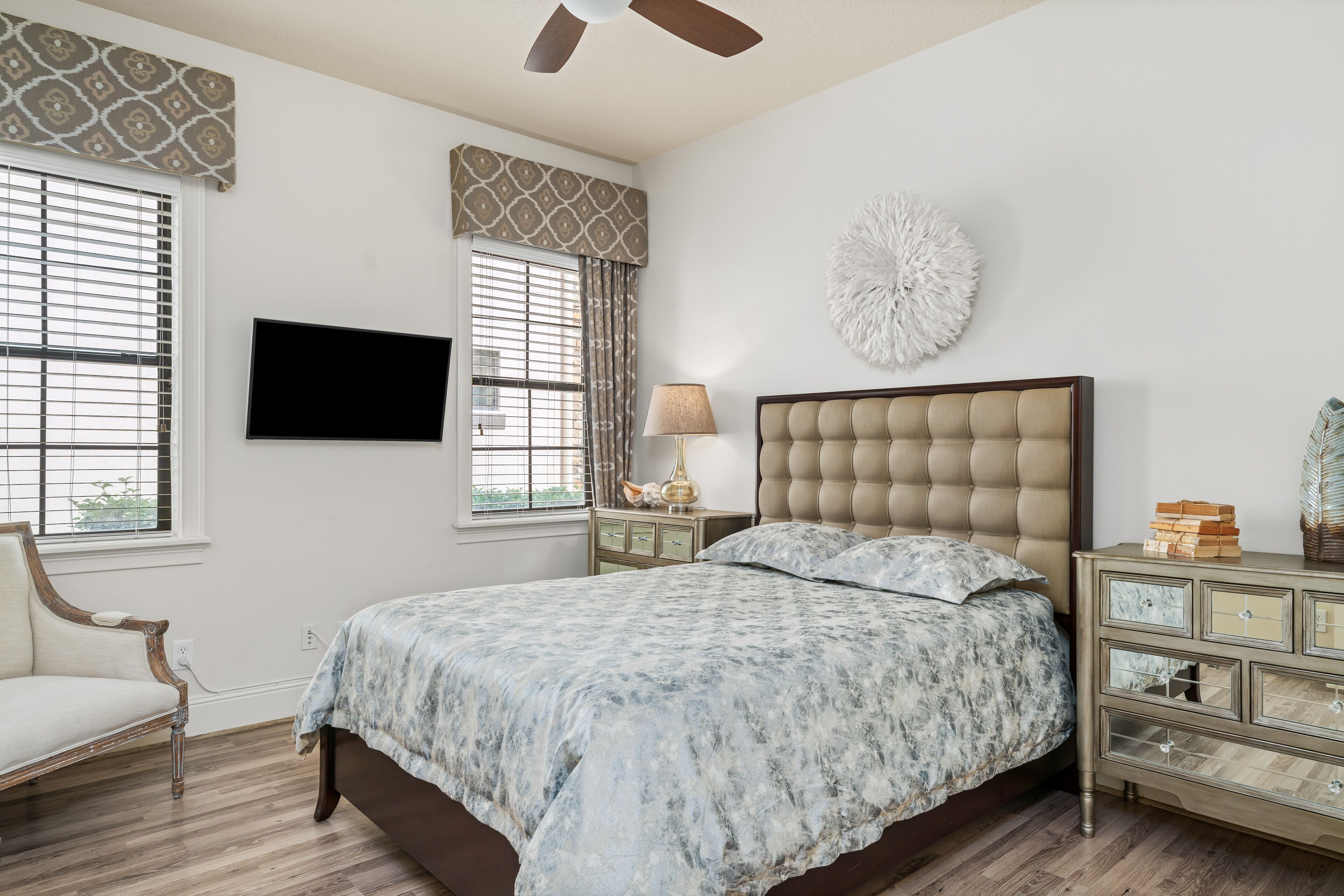 Un dormitorio con una cama grande, televisión montada en la pared, ventilador de techo y un adorno de pared decorativo.