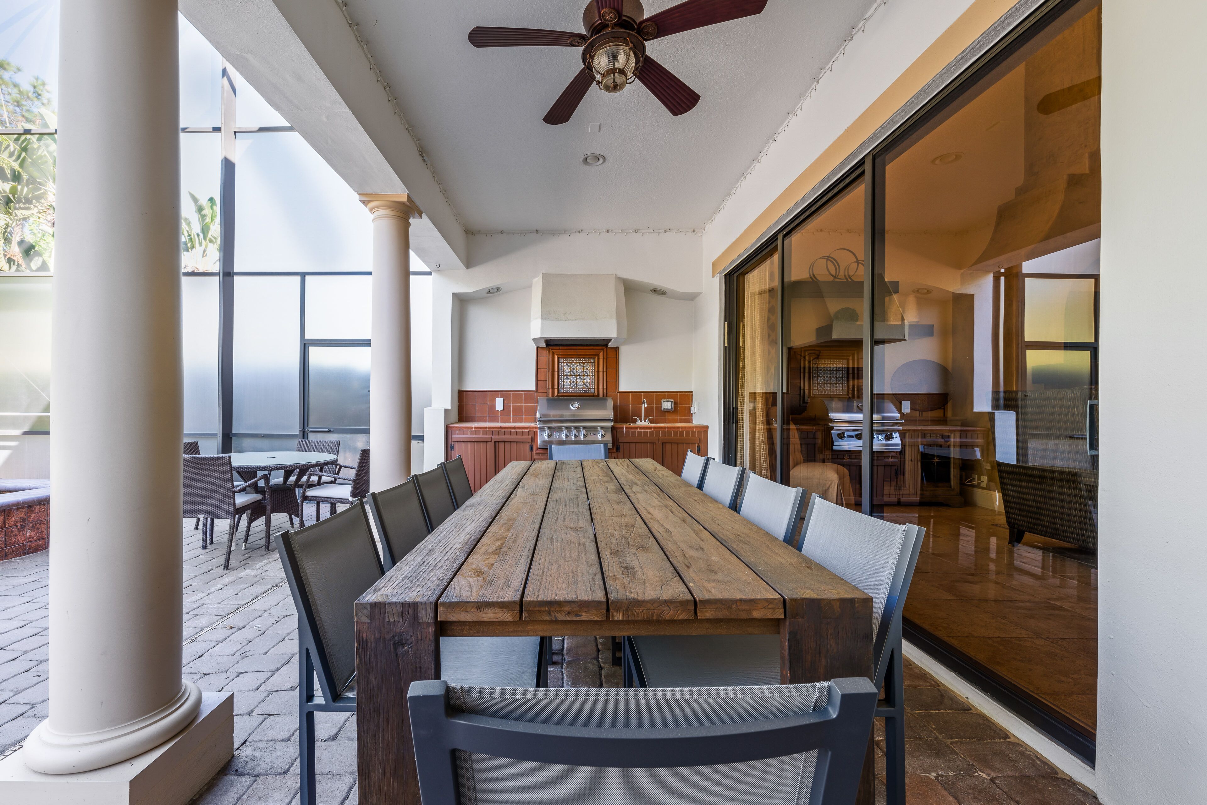 A spacious patio with a wooden table and chairs, a ceiling fan, and a view of the kitchen and living area through glass doors.