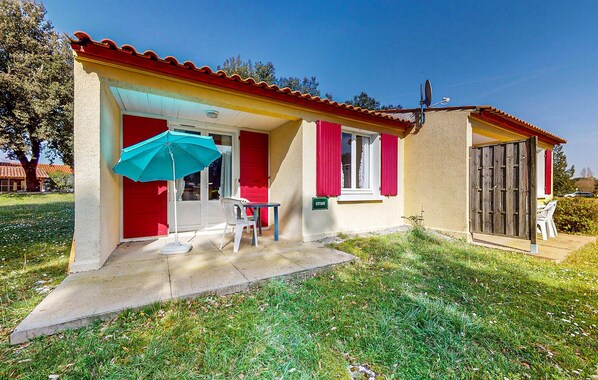 Una casa pequeña de un piso con puerta y ventanas rojas, un patio con mesa y sillas, y un paraguas azul.