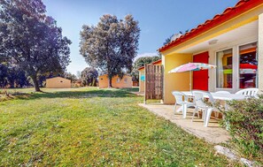 Un espacio exterior soleado con césped, sillas de plástico blanco y una mesa bajo un paraguas colorido.