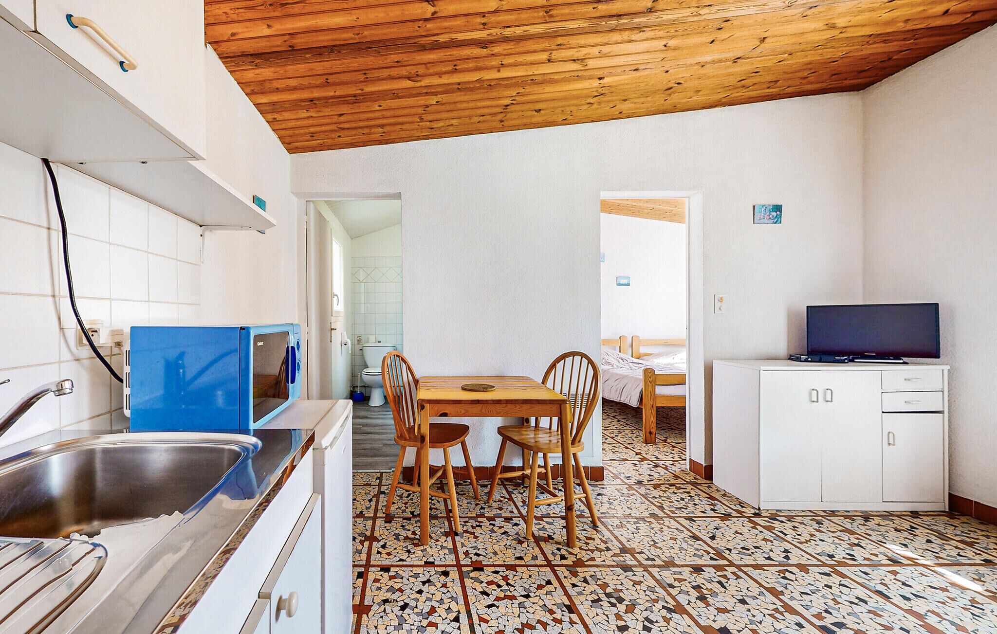 Een compacte keuken met houten plafond, een eethoek met tafel en stoelen, een wit kastje met een televisie, en een betegelde vloer.
