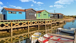 En träbrygga med färgglada trähus, båtar förtöjda vid bryggans ände och stilla vatten som speglar byggnaderna.