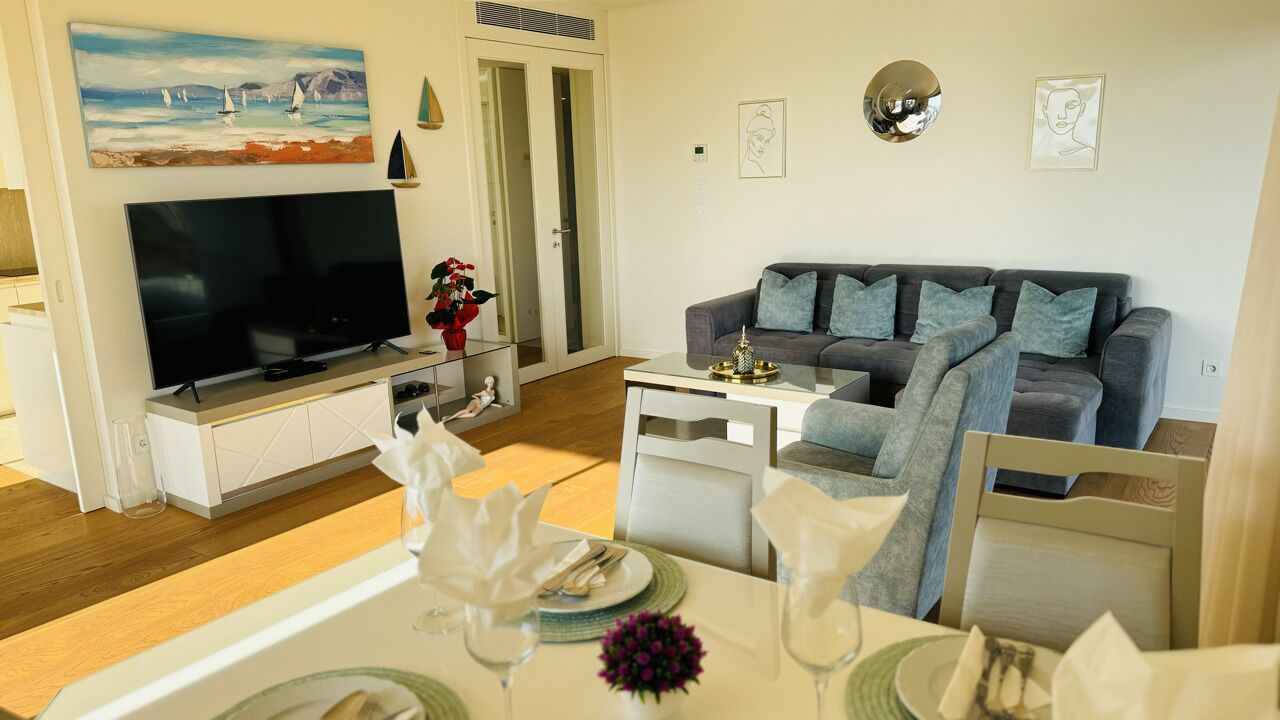 A modern living room with a flat-screen TV, grey sofa, and a dining area set with a table, chairs, and a vase of flowers.