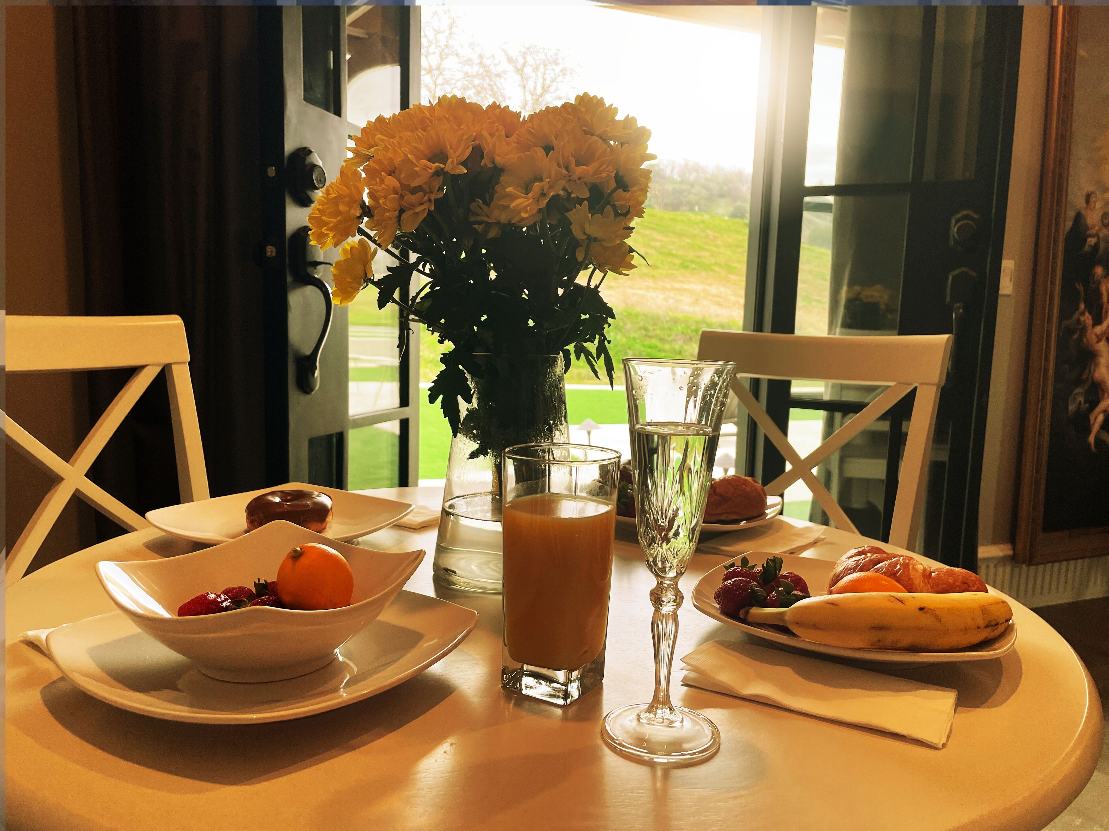 A dining table with a bowl of strawberries, a glass of orange juice, a glass of water, a banana, and a plate of croissants. There is a vase with flowers and a painting on the wall.