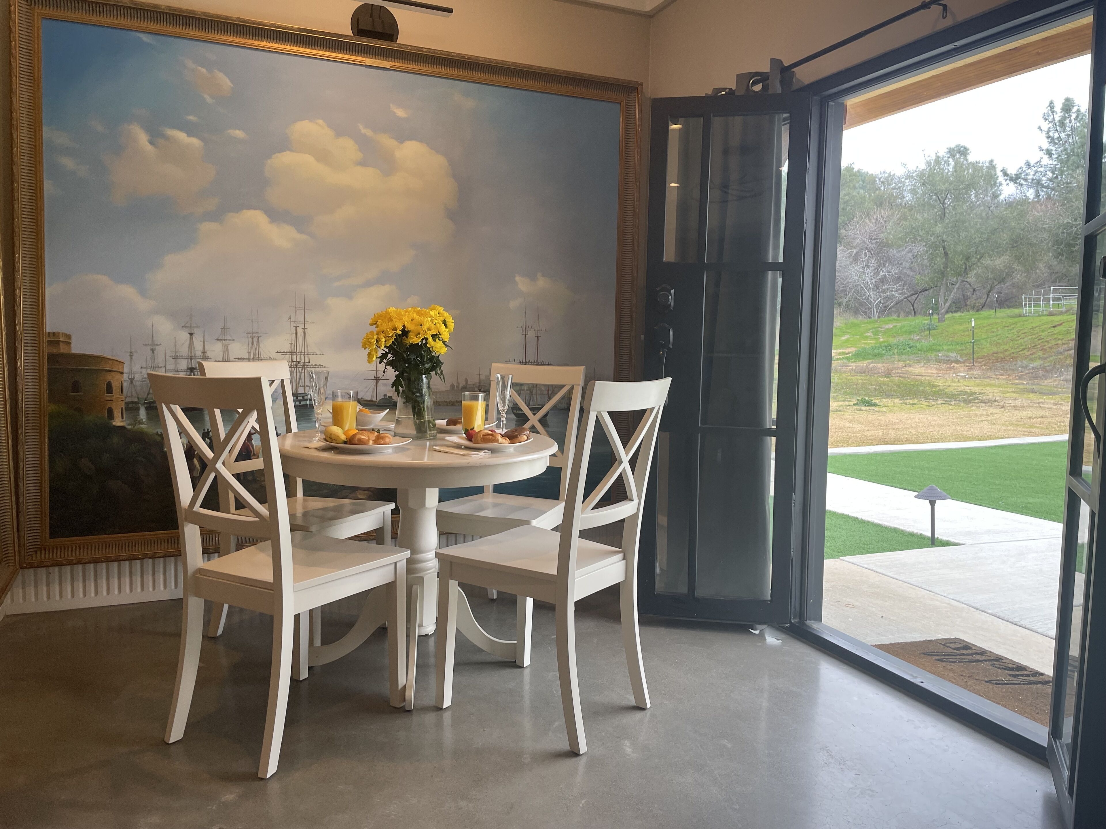Un coin repas avec une table ronde, des chaises blanches, un tableau représentant des navires accroché au mur et une vue sur l’extérieur.