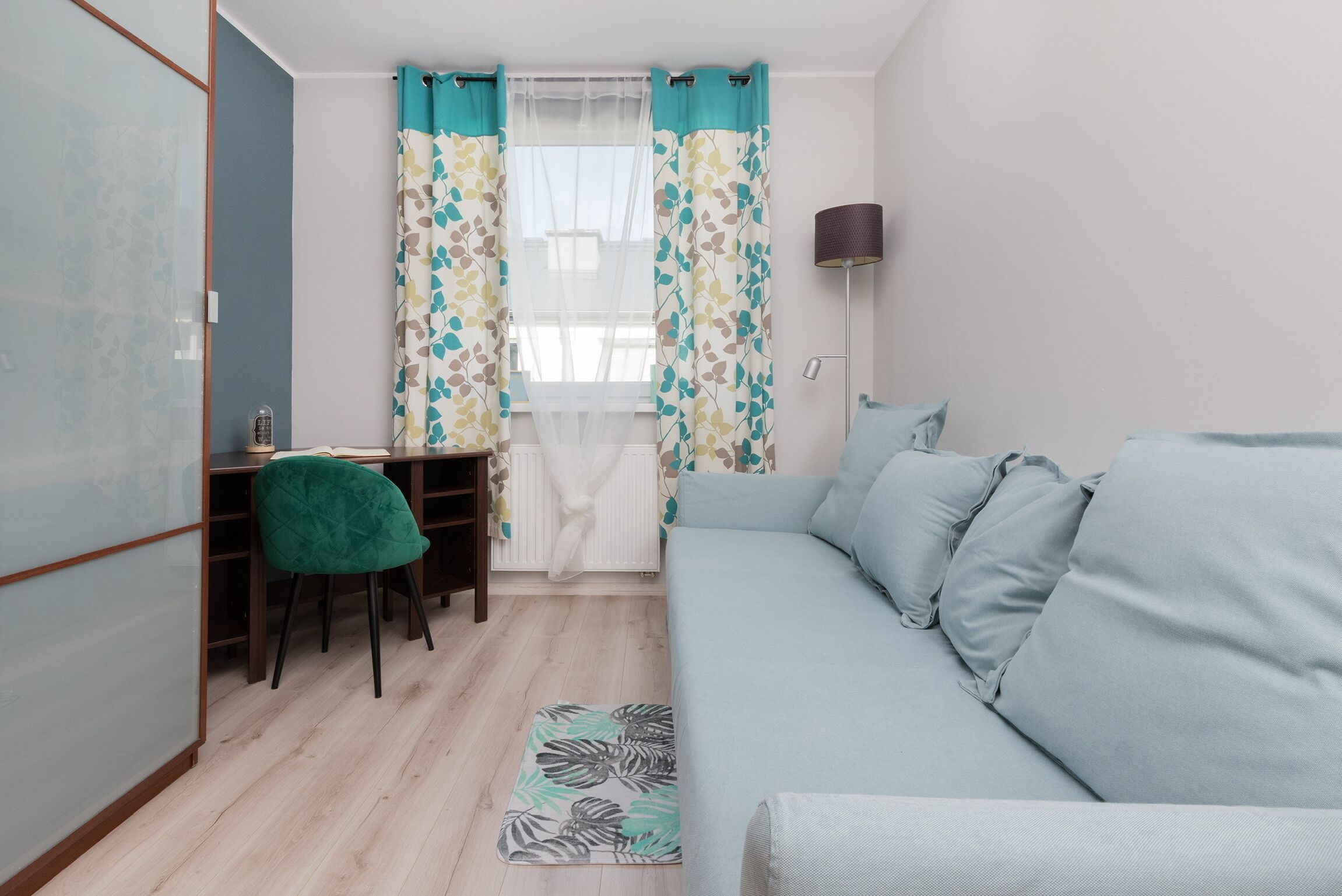 Une petite pièce bien éclairée, avec un canapé bleu clair, un fauteuil vert, un bureau et une fenêtre ornée de rideaux à motifs.