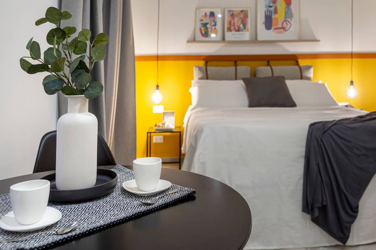 A hotel room with a bed, bedside table, and a vase with green leaves.