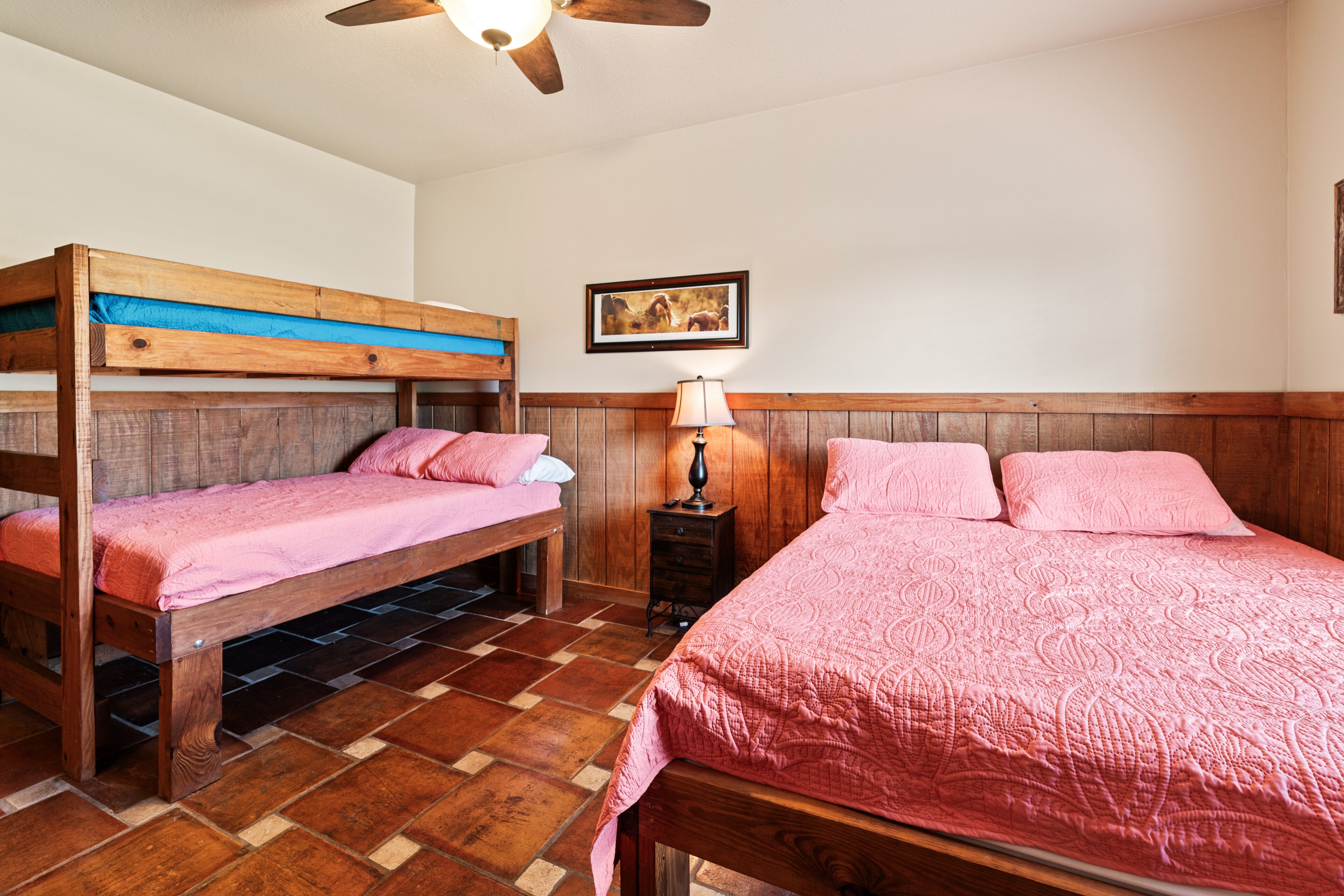 Un dormitorio con dos literas y una cama individual, suelo de madera, ventilador de techo y un cuadro en la pared.
