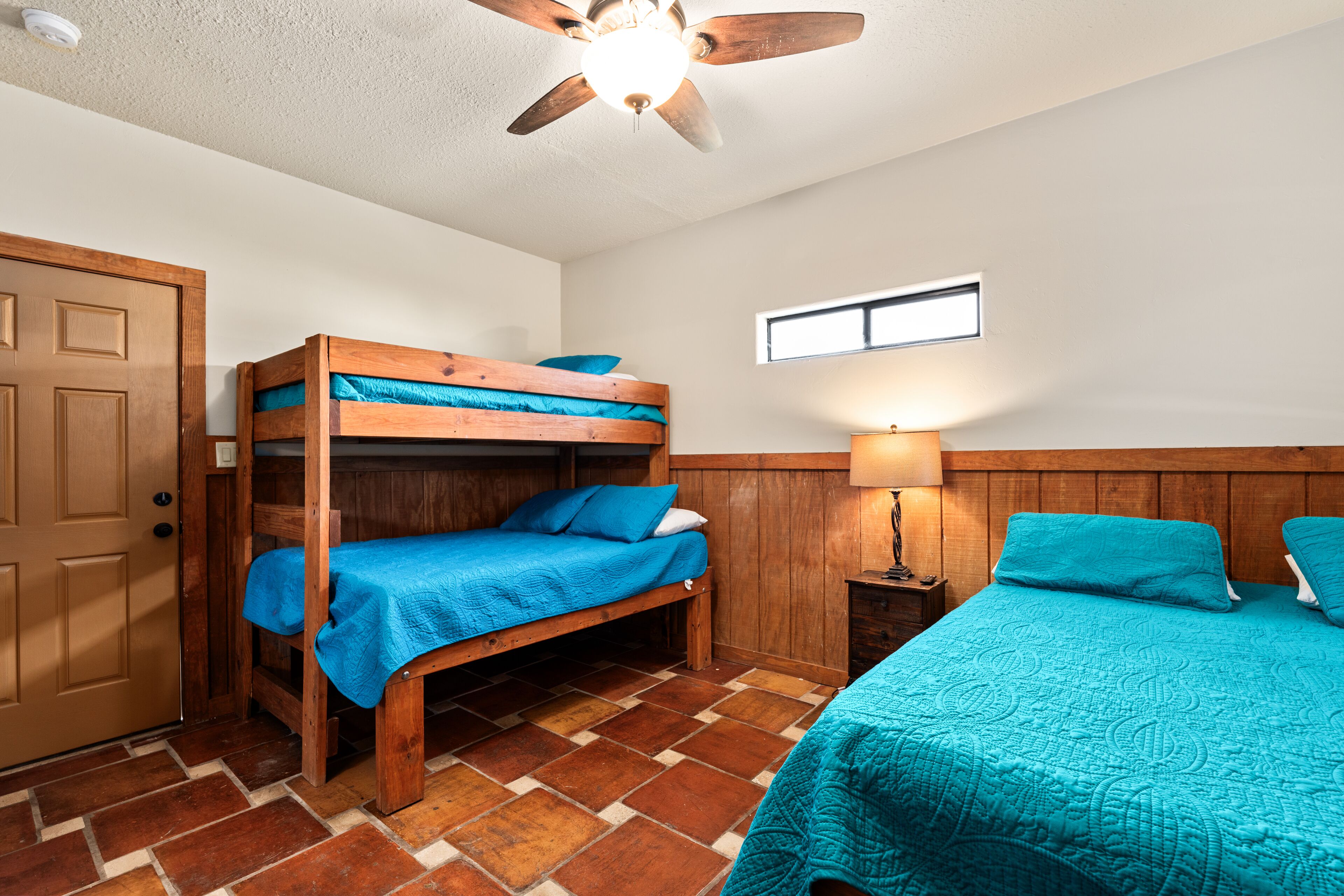 A room with a bunk bed, a single bed, a wooden door, a window, and a ceiling fan.