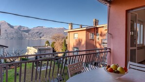 A balcony with a table and a bowl of fruit, overlooking a lake and mountains.
