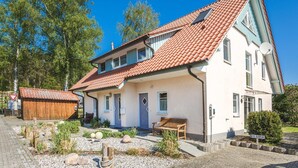 Une maison à deux étages, avec un toit recouvert de tuiles rouges, un porche à l’avant doté d’un banc, et une petite construction en bois située sur la gauche.