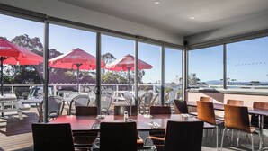 An outdoor dining area with tables and chairs, red umbrellas, and large windows offering a view of the surrounding landscape. - Bridport Bay Inn (Bridport)