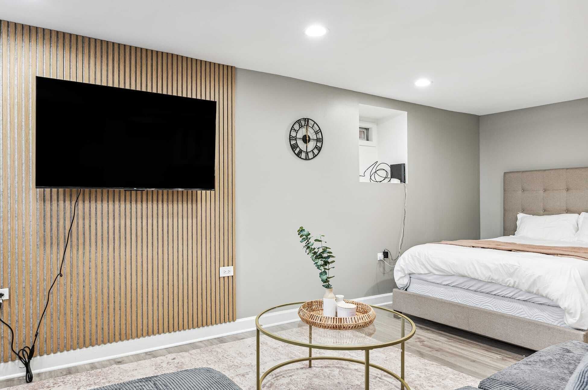 A modern bedroom with a flat-screen TV mounted on a wooden panel, a round table with a woven basket, a bed with white bedding, and a wall clock.