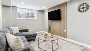 A modern living room with a grey sofa, a round coffee table, and a flat-screen TV mounted on a wooden panel wall.