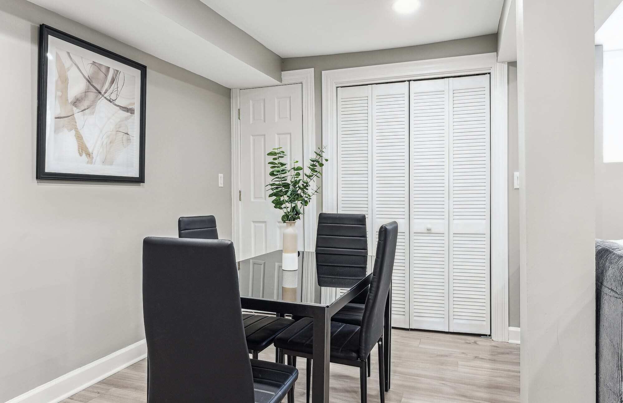A dining area with a glass table and black chairs, a framed abstract painting, a white door, and a white closet with louvered doors.