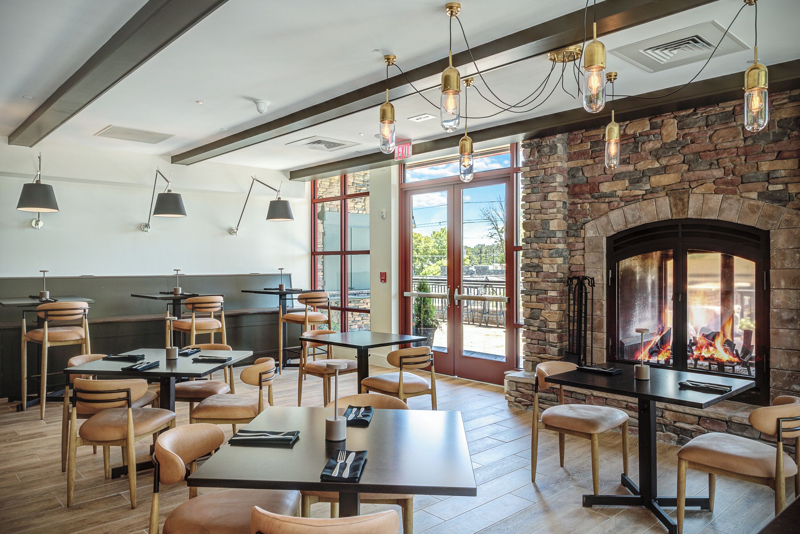 A restaurant with wooden tables and chairs, hanging pendant lights, and a stone fireplace with a visible fire.