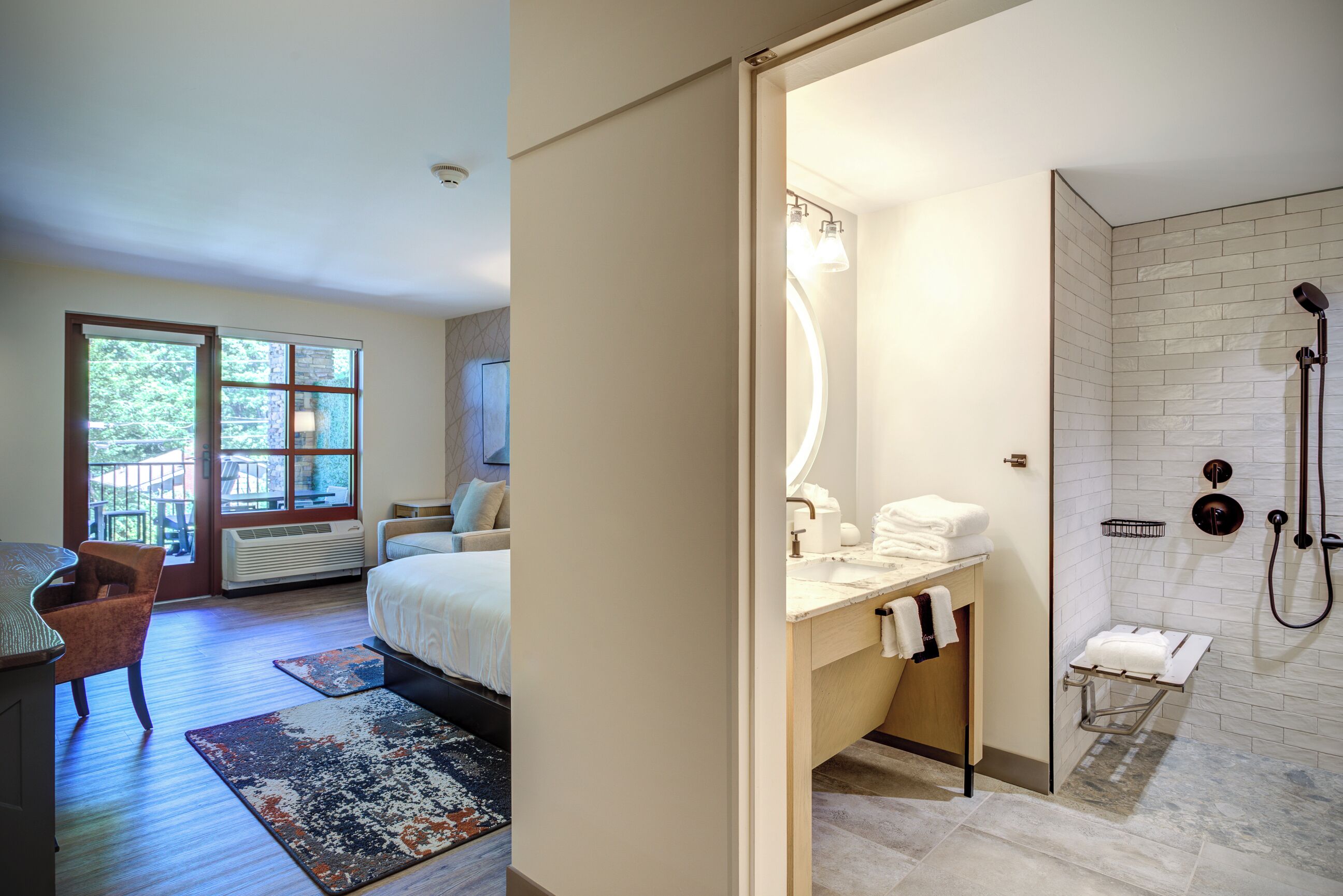 A hotel room with a bed, a desk, a chair, a bathroom with a shower, and a view of the outdoors.