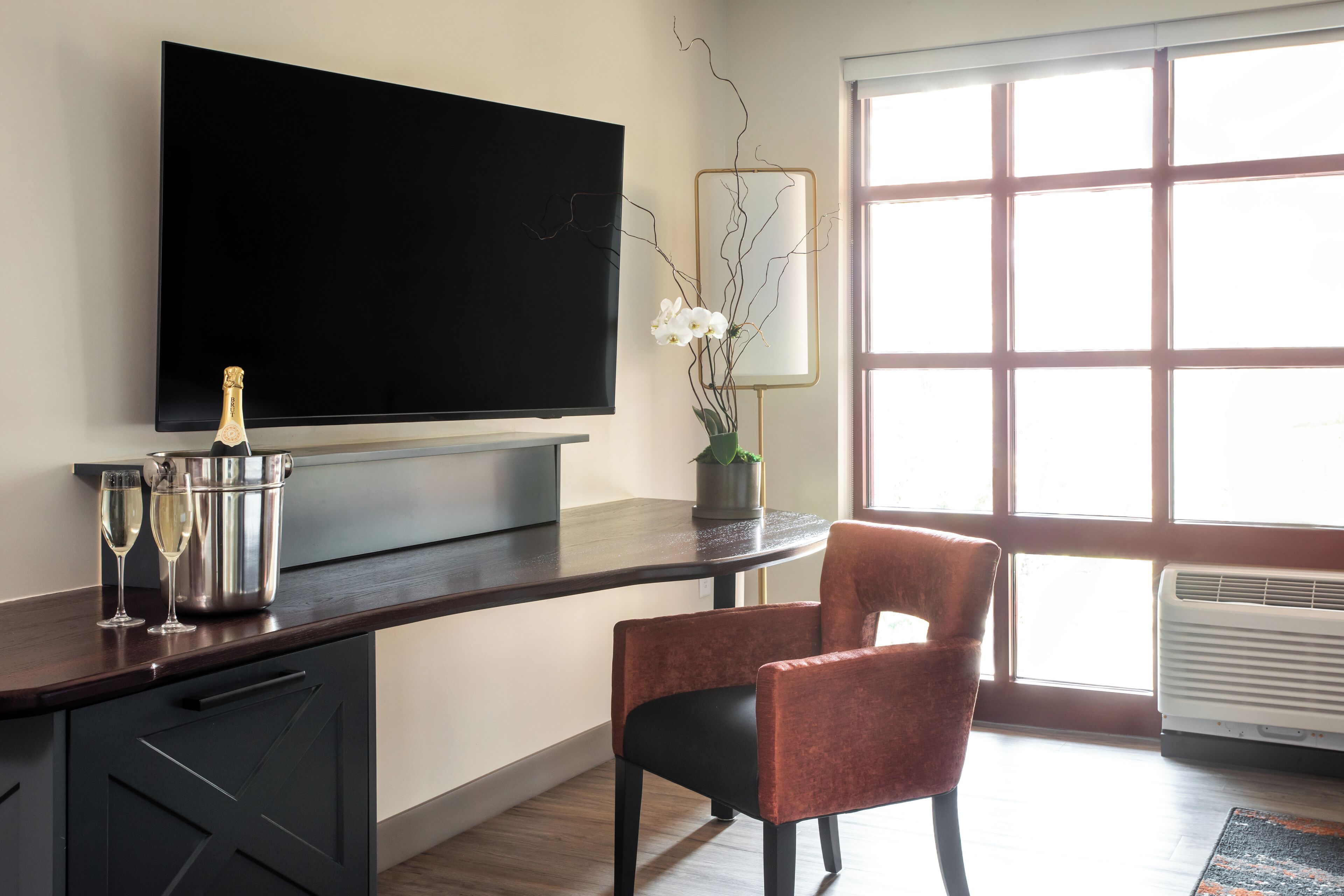 A modern living room with a flat-screen TV mounted on the wall, a dark wooden coffee table, a red upholstered chair, and a large window providing natural light.