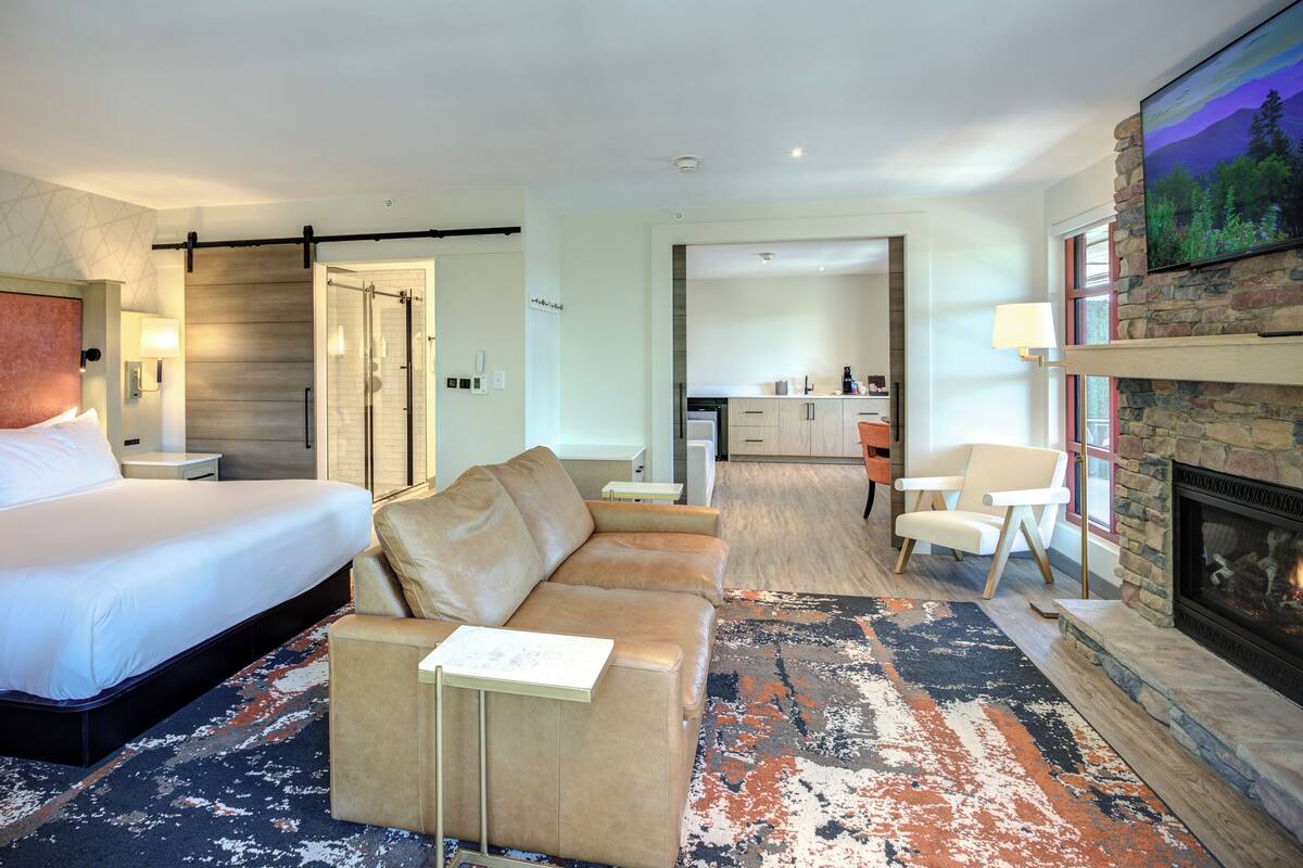 A hotel room with a bed, a sofa, a small table, a fireplace, and a kitchen area in the background.