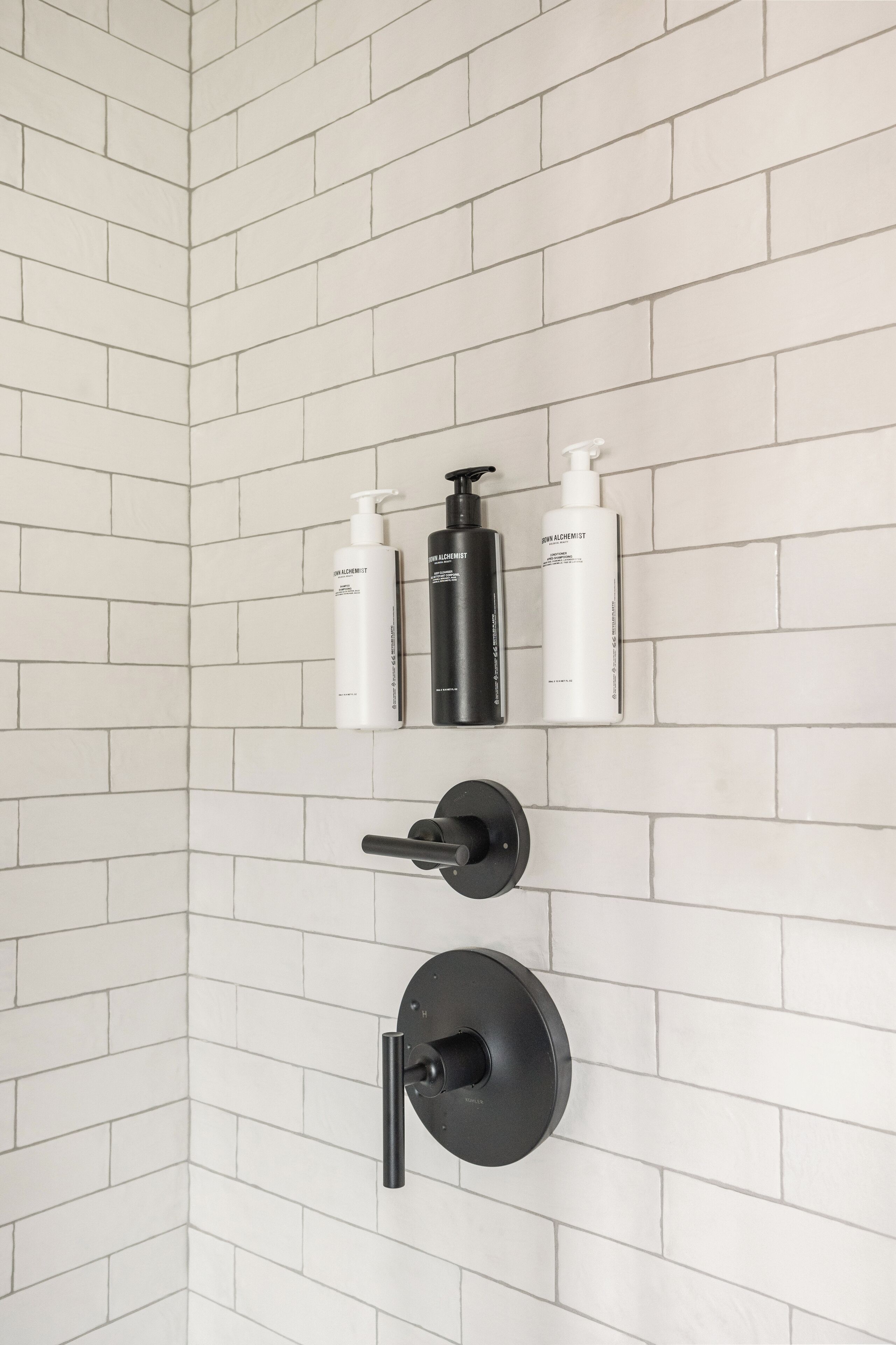 A corner of a bathroom with white tiled walls, a black faucet, and three bottles of skincare products.