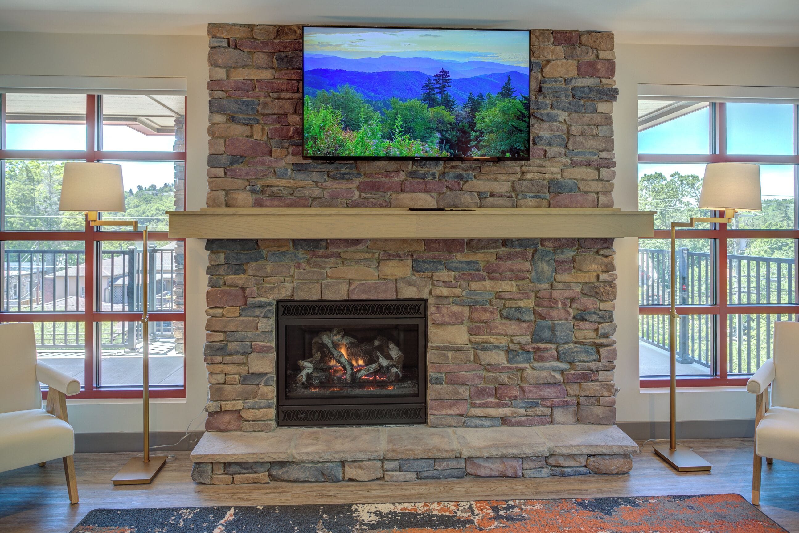 A stone fireplace with a TV mounted above it, two lamps on either side, and a view of the outdoors through large windows.