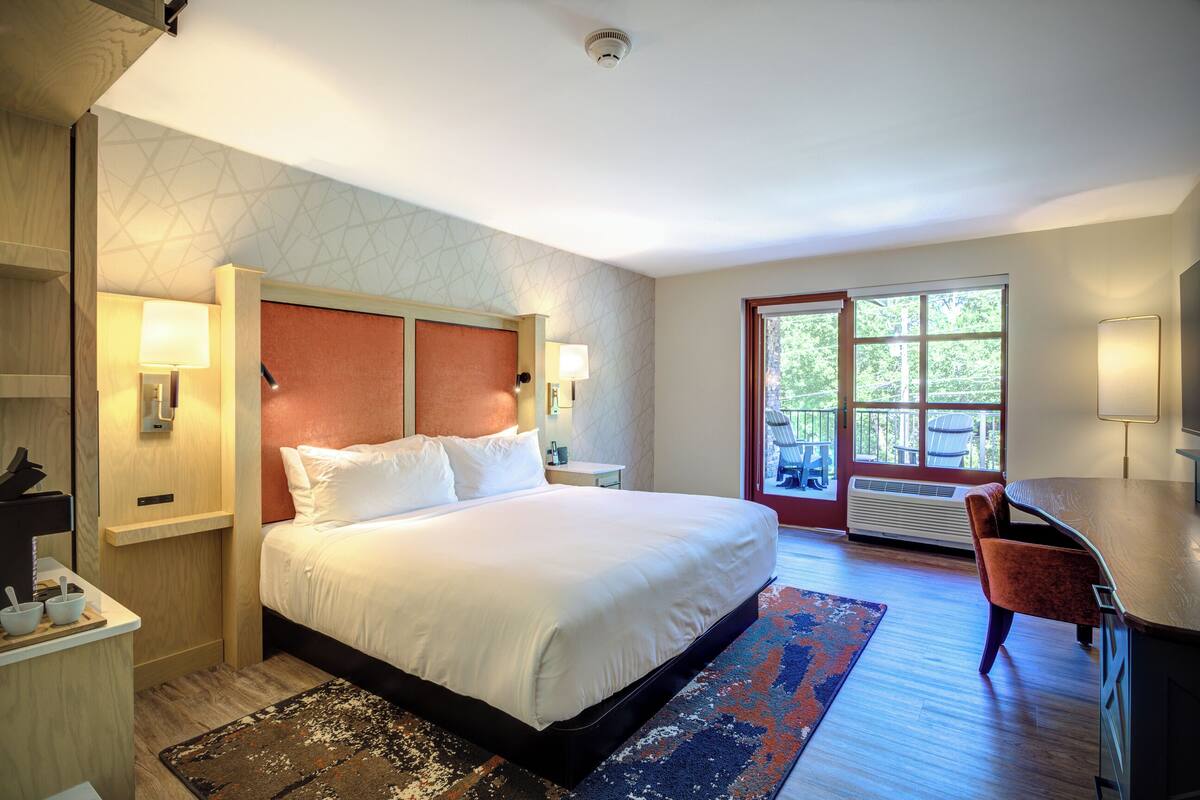 A hotel room with a large bed, a desk, a chair, and a view of the outdoors through a glass door.