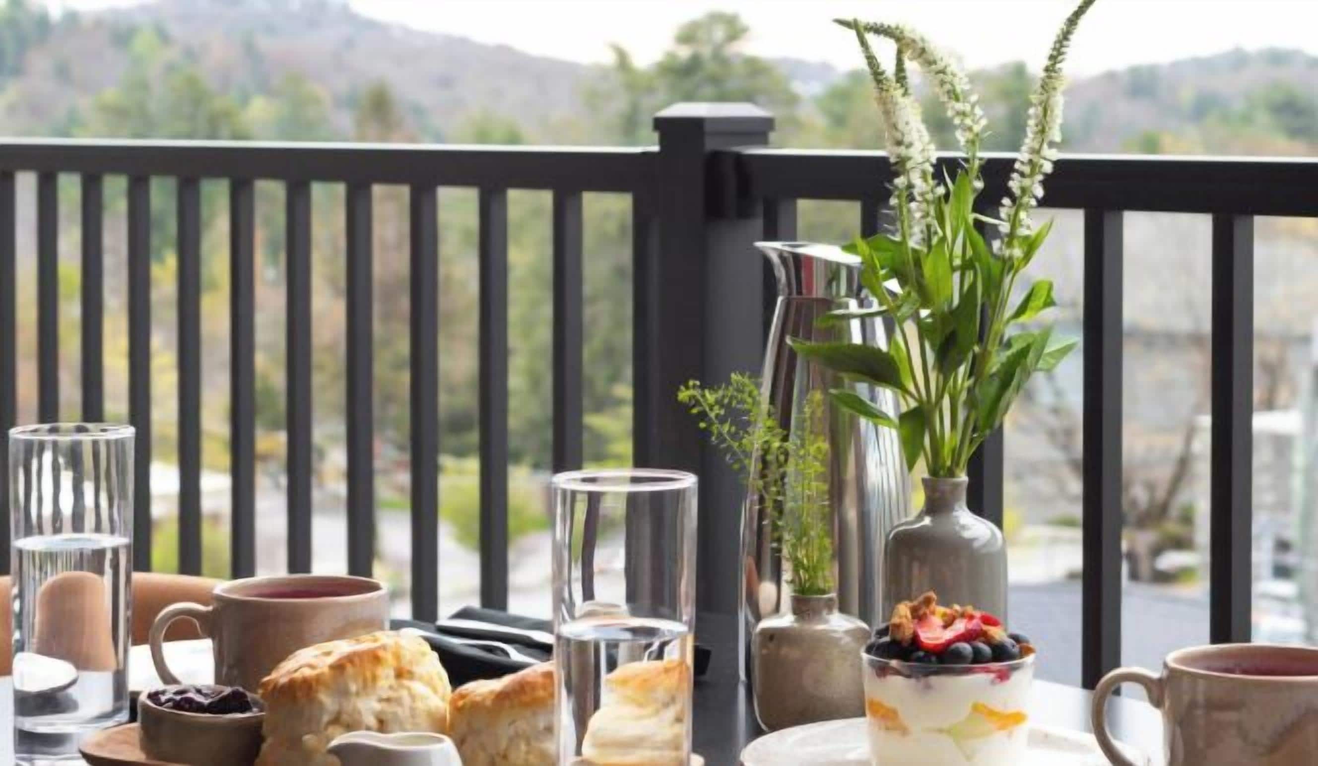 A breakfast setup with pastries, a glass of water, a mug, and a vase with flowers on a balcony with a scenic view.
