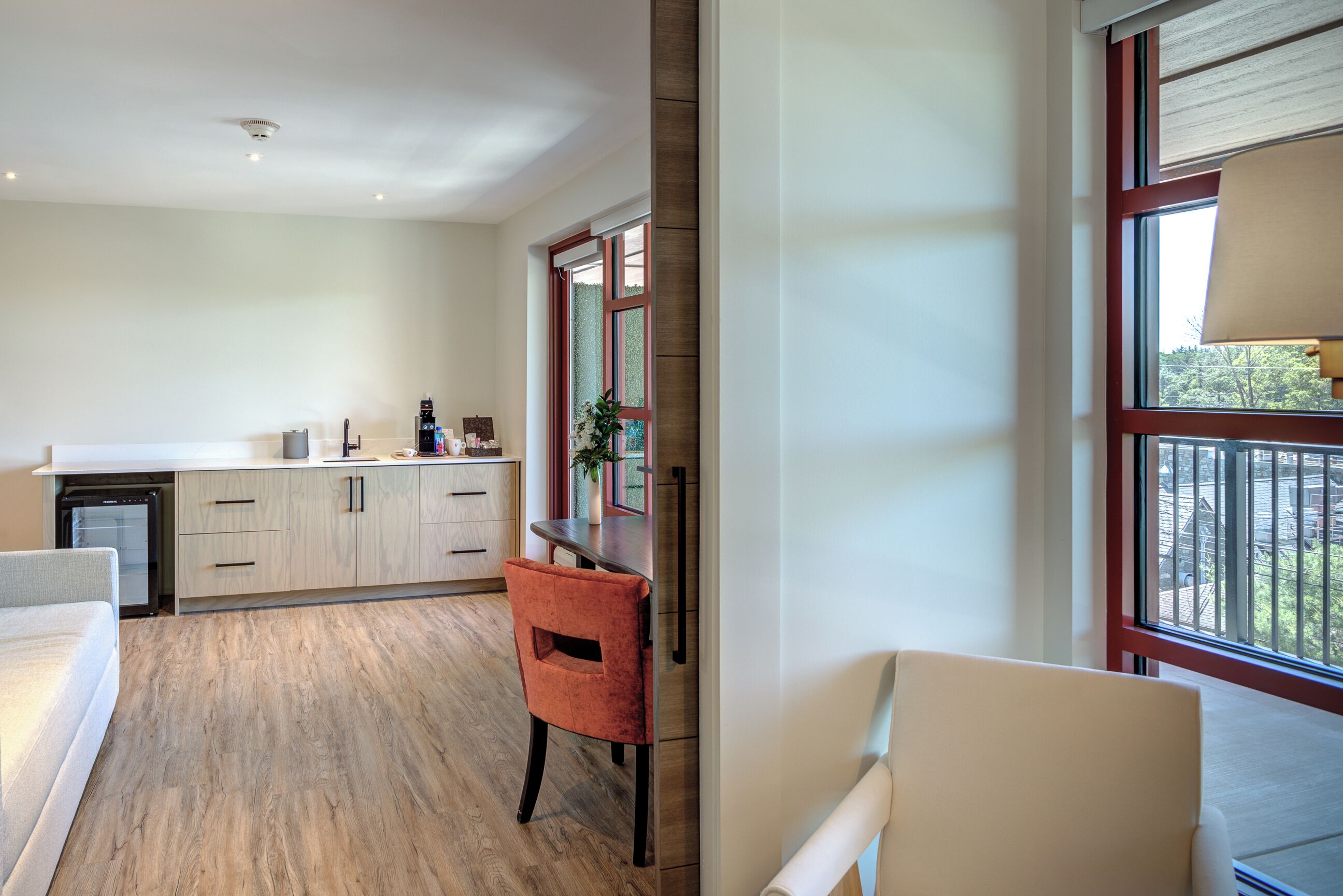 A modern kitchen with wooden flooring, a dining area with a red chair, and a view of the outdoors through large windows.