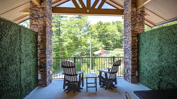 Un patio cubierto con pilares de piedra, dos sillas tipo Adirondack, una mesita y vistas del entorno verde.