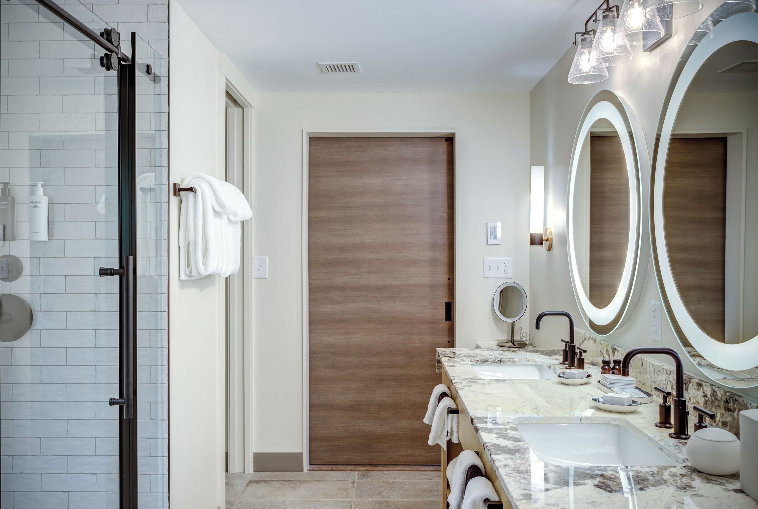 A modern bathroom with a glass shower enclosure, a wooden door, and a vanity with two sinks and large mirrors.