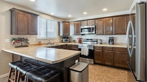 A kitchen with dark wooden cabinets, a stainless steel refrigerator, a microwave, an oven, and a coffee maker. There is a bar stool at the island and a trash can. - Homestead Haven | Sleeps 14 | 2 Car Garage (Clearfield)