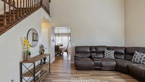 A living room with a brown leather sectional sofa, a wooden console table with a mirror, and a staircase with wooden railings. - Homestead Haven | Sleeps 14 | 2 Car Garage (Clearfield)