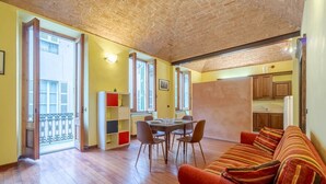A room with a wooden floor, a dining table with chairs, a sofa, and a kitchen area visible through an open door.