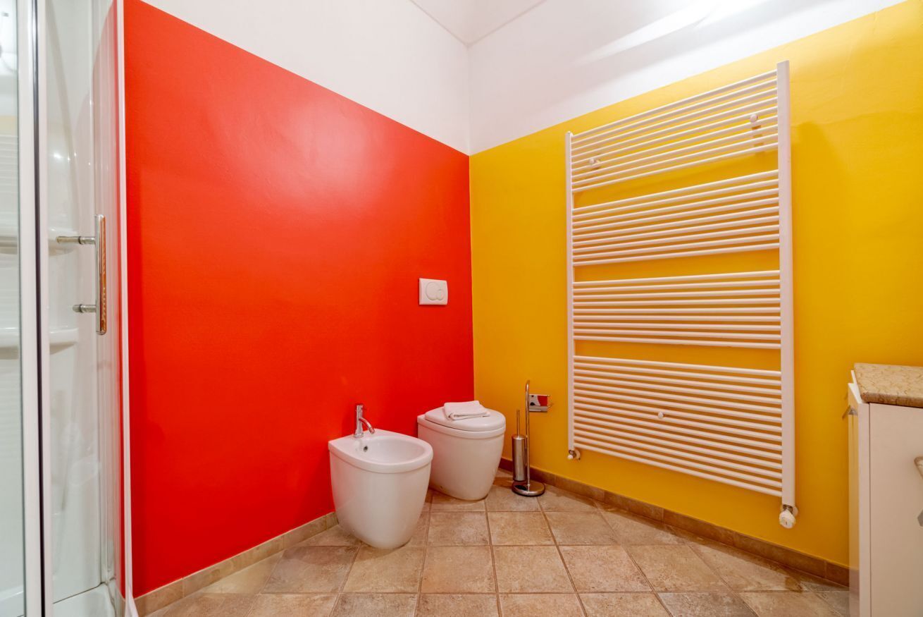 A bathroom with red and yellow walls, a white radiator, two toilets, and a white sink.