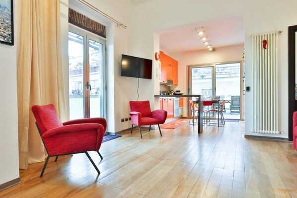 A modern living room with a flat-screen TV, red chairs, a dining area, and a kitchen visible through an open door.