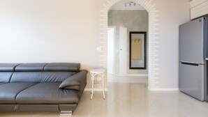 Una sala de estar moderna con un sofá gris, un taburete blanco pequeño y una zona de cocina con un refrigerador.